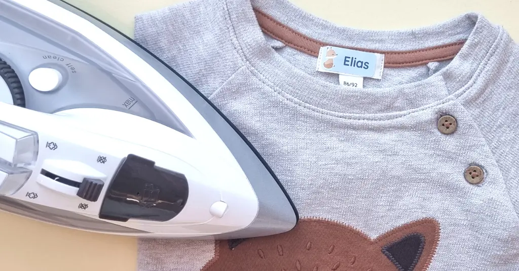 Bügeletikett mit Fuchsmotiv auf einem grauen Babyshirt, daneben ein Bügeleisen