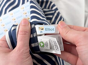 Hand bringt einen blauen Namensaufkleber mit Löwenmotiv und dem Namen Elliot auf einem Etikett in einem gestreiften Shirt an