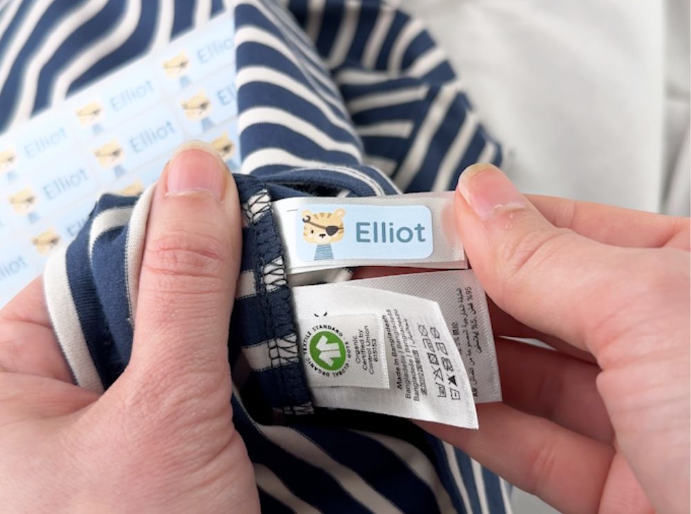 Hand bringt einen blauen Namensaufkleber mit Löwenmotiv und dem Namen Elliot auf einem Etikett in einem gestreiften Shirt an