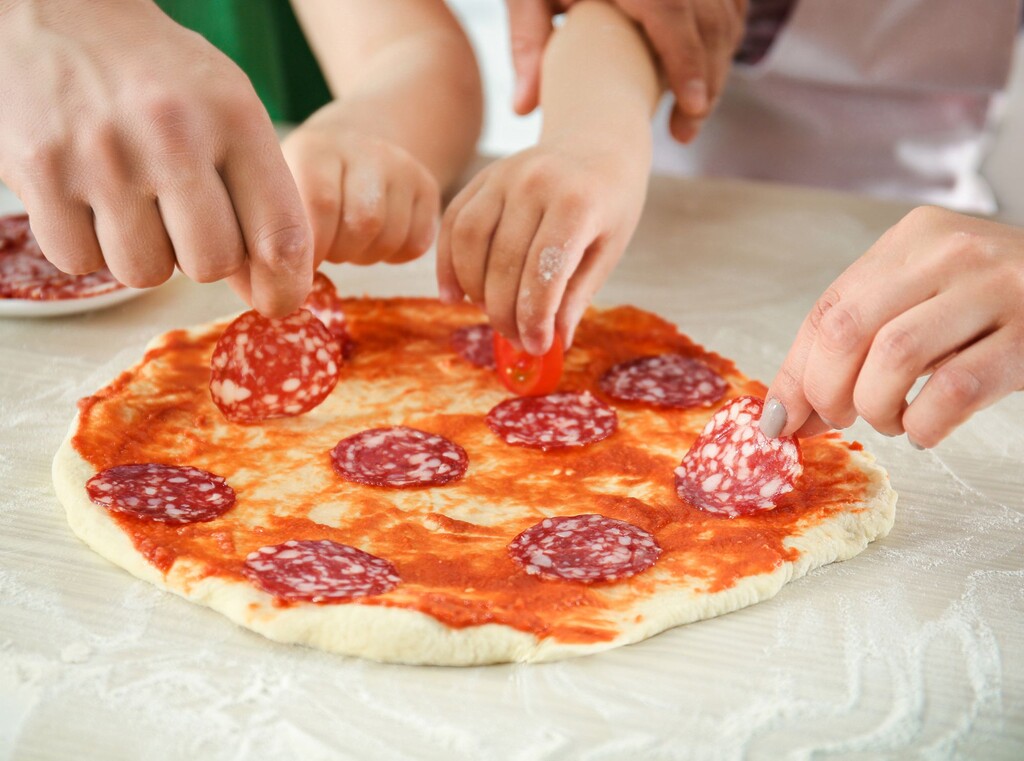 Pizza backen mit kindern: einfache rezepte, mini-pizzen und schneller pizzateig zum selbermachen 4 Kinder- und erwachsenenhände belegen gemeinsam eine pizza