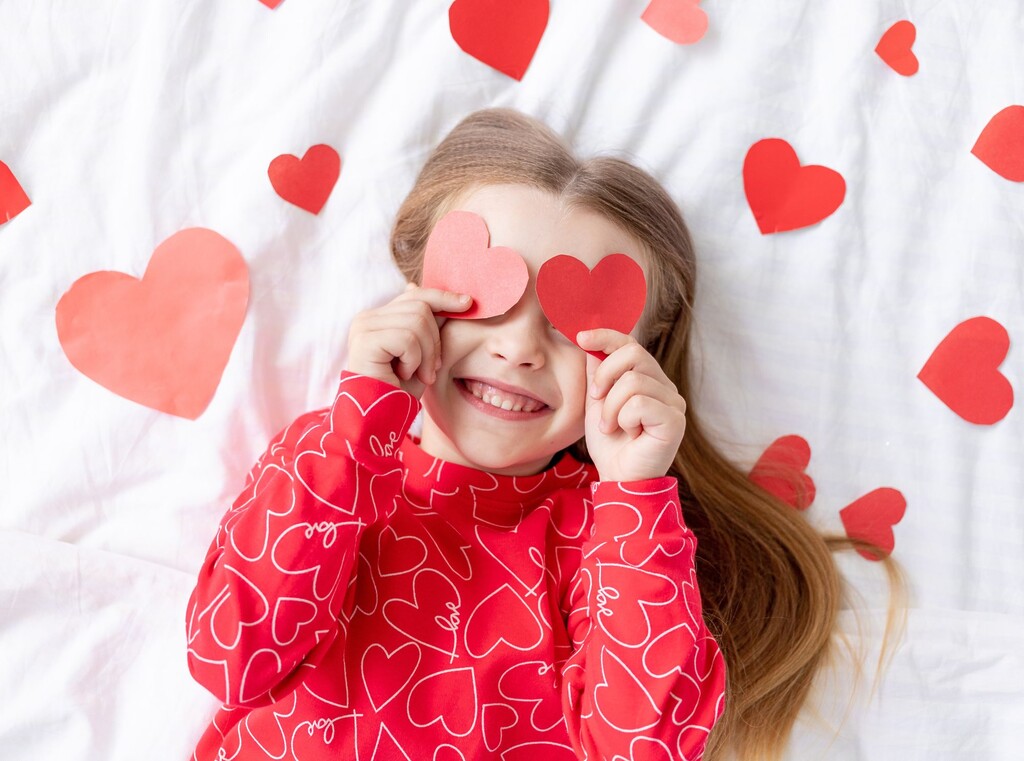 Glücklich lächelndes Kind mit Herzchen-Pulli hält sich zwei gebastelte rote Herzen vor die Augen