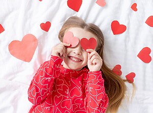 Glücklich lächelndes Kind mit Herzchen-Pulli hält sich zwei gebastelte rote Herzen vor die Augen