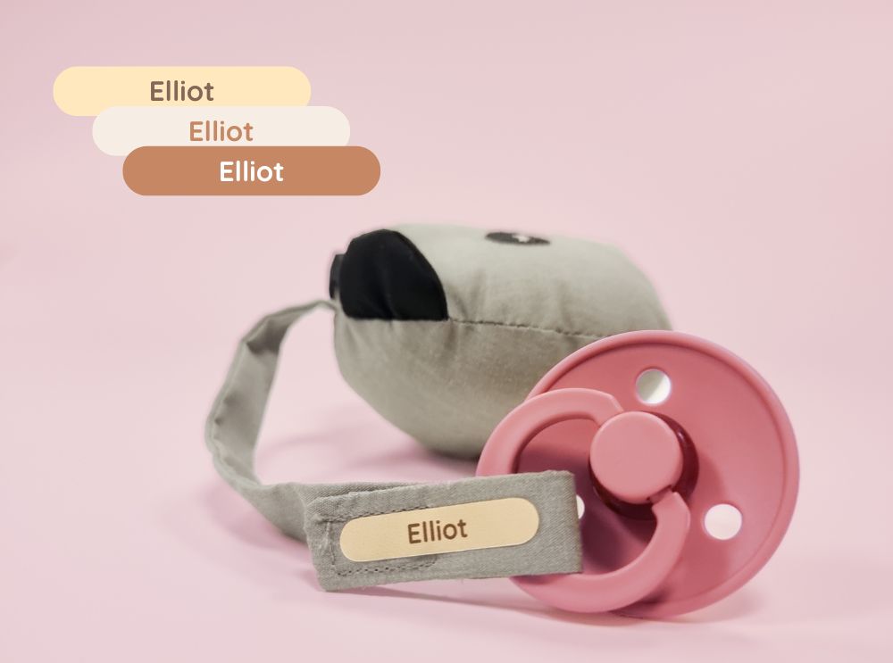 Pink pacifier with a personalized name label reading 'elliot' on a fabric strap.