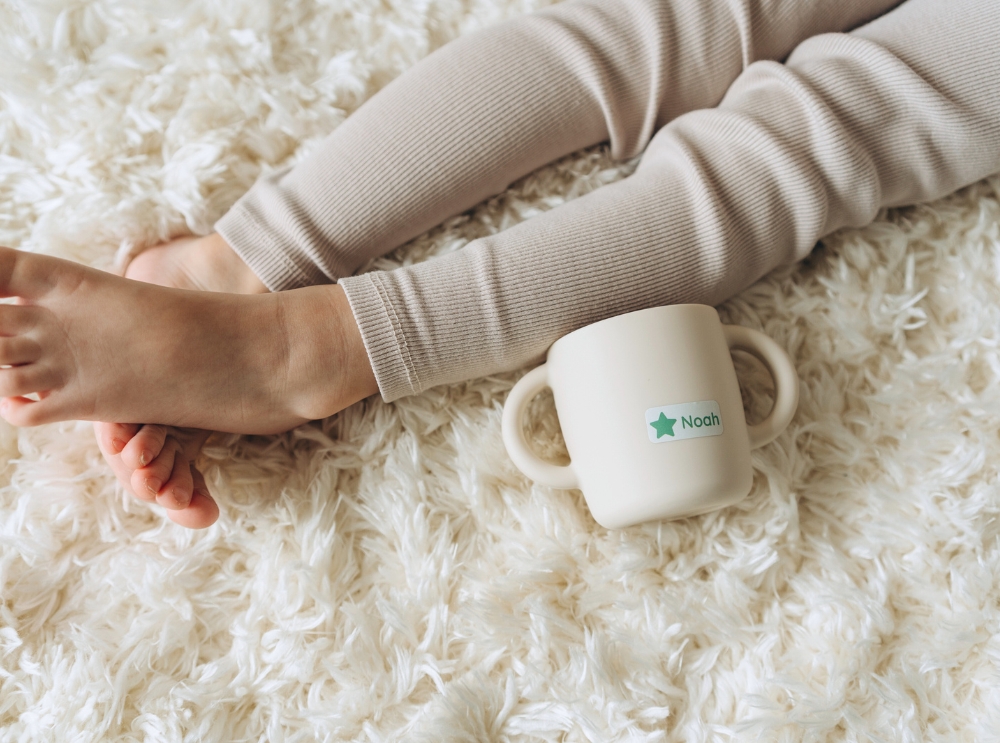 Toddler cup with a personalized name label on a soft, fluffy rug next to a child's legs.