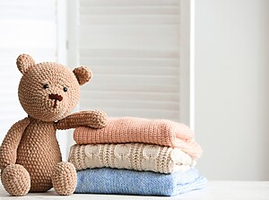Gestrickter Teddybär neben einem Stapel gefalteter Babykleidung in sanften Pastellfarben