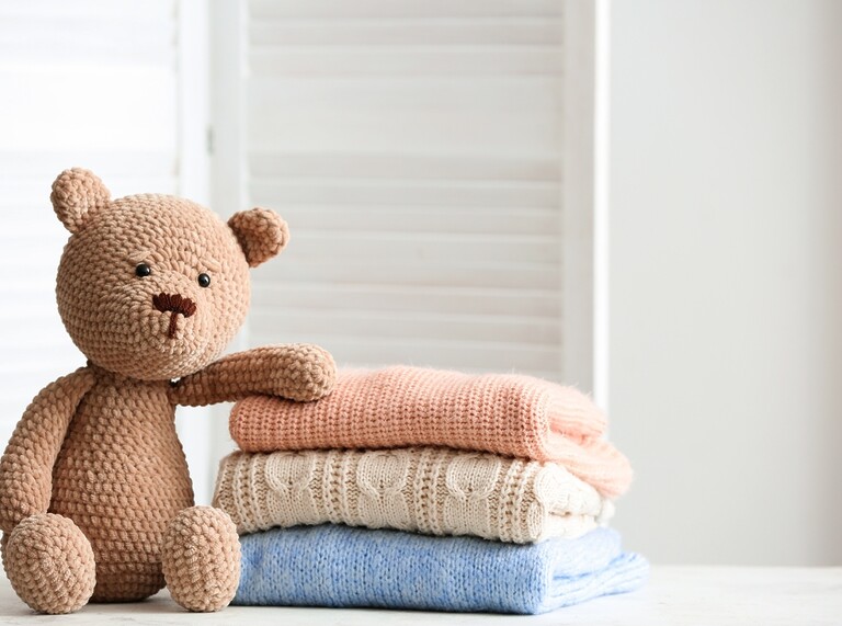 Gestrickter Teddybär neben einem Stapel gefalteter Babykleidung in sanften Pastellfarben