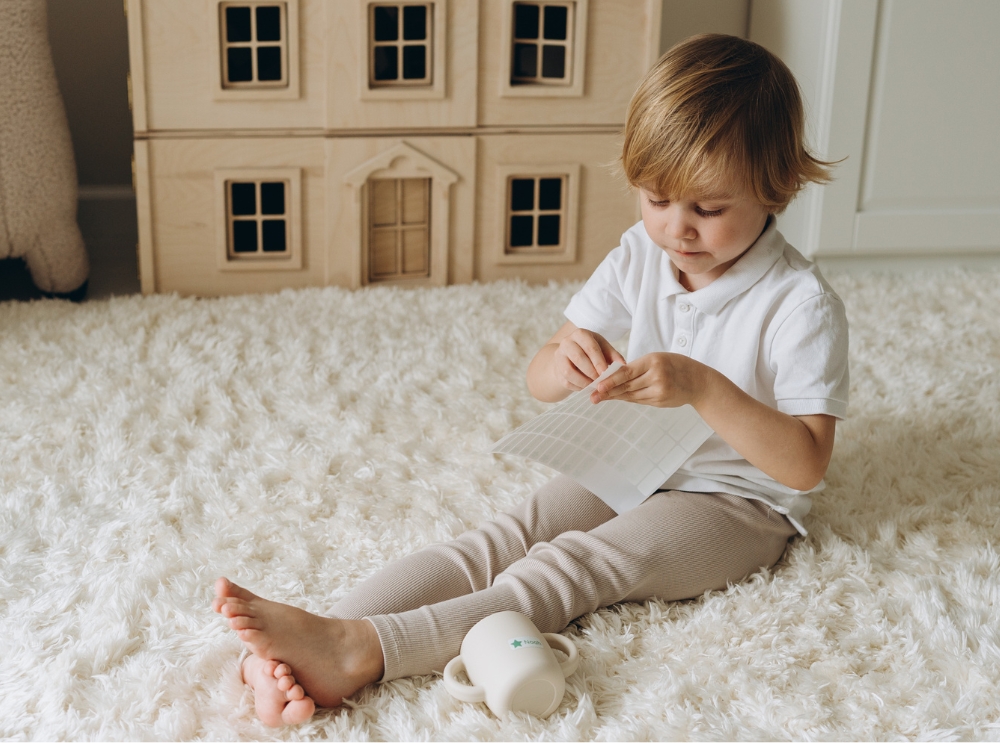 Kind sitzt auf einem weichen Teppich und löst einen Namensaufkleber von einem Etikettenbogen im gemütlichen Spielzimmer