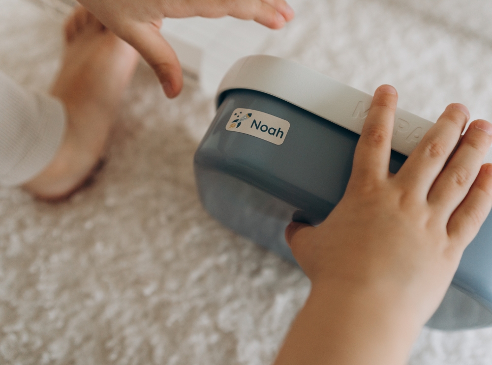 Nahaufnahme von kinderhänden, die eine blaue brotdose mit einem personalisierten namensaufkleber mit raketenmotiv und dem namen noah halten