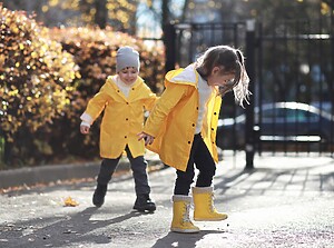 Kinder in Regenmänteln und Gummistiefeln spielen in Herbstlaub an einem sonnigen Tag