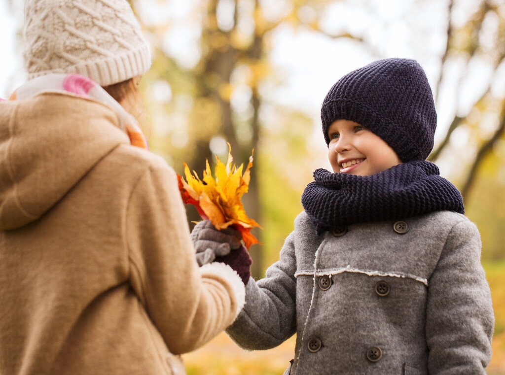 Herbstkleidung für kinder: so bleiben deine kinder im herbstwetter warm und trocken 3 Kinder in wintermänteln und mützen tauschen herbstblätter in einem park aus.